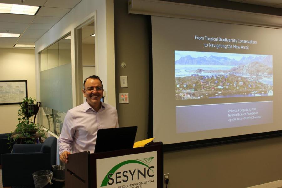 Roberto Delgado standing behind a podium in front of a presentation projected on a screen