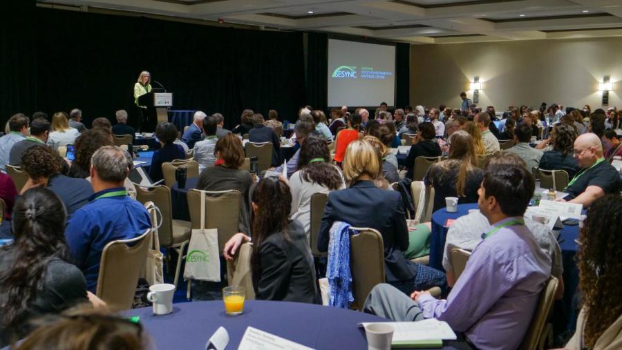 People seated at tables listening to a speaker at the symposium