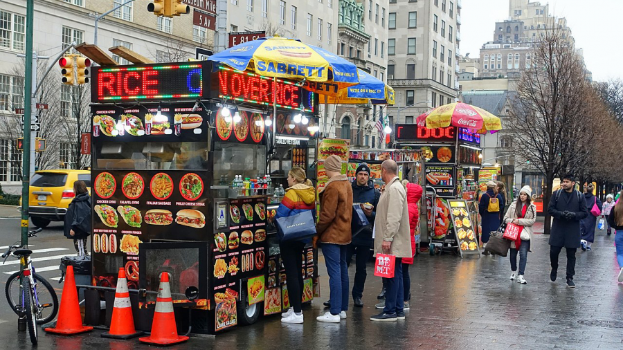 Several food trucks/stands lined along a New York City street with people lined up to make a purchase