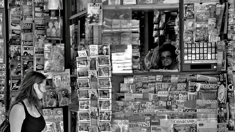 A black and white photo of a woman walking past a newsstand in Seville, Spain, looking at the newspapers and magazines, while a woman working behind the stand looks on through the window